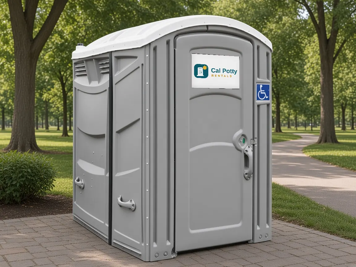 A grey ADA-accessible portable restroom with a wheelchair sign on a paved path in a tree-lined park