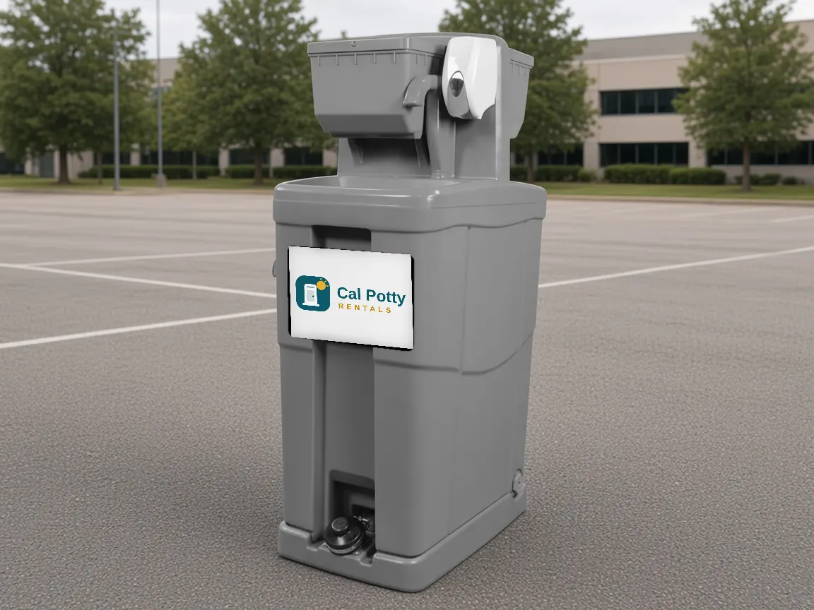 A grey handwashing station in an empty parking lot with trees and an office building behind